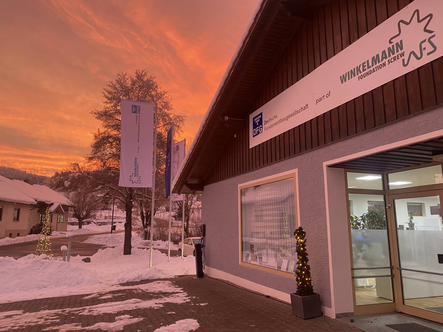 Schneebedecktes Gebäude mit dem Schild „Winkelmann“, beleuchtet von einem leuchtend orangen Sonnenuntergang. Draußen sind Fahnen und Bäume zu sehen.