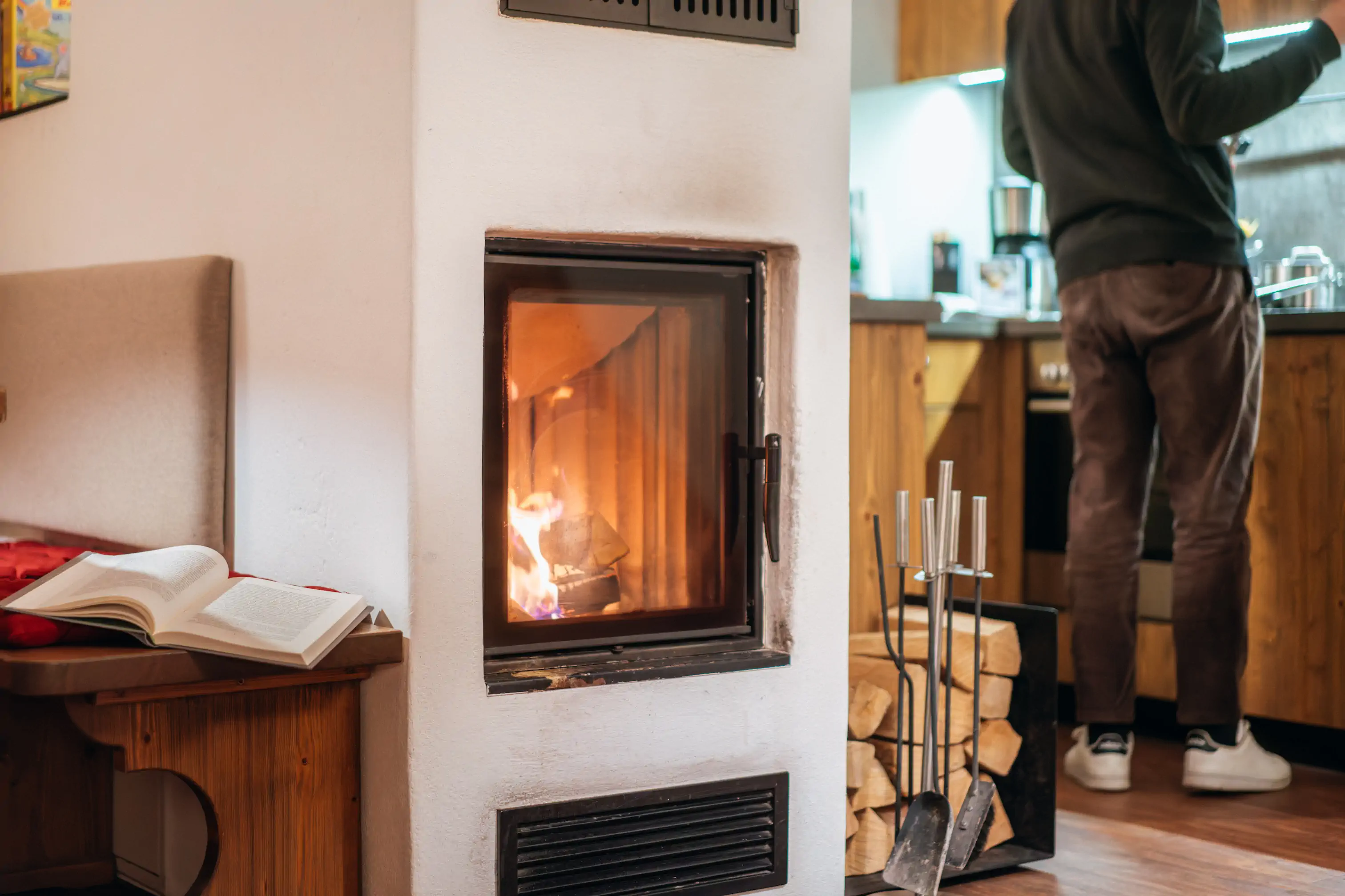 Gemütliches Wohnzimmer in einem Ferienhaus im Harz mit brennendem Kamin und einem aufgeschlagenen Buch auf einer Sitzbank sowie einer Person, die in einer Küche mit Holzschränken und Küchengeräten kocht.