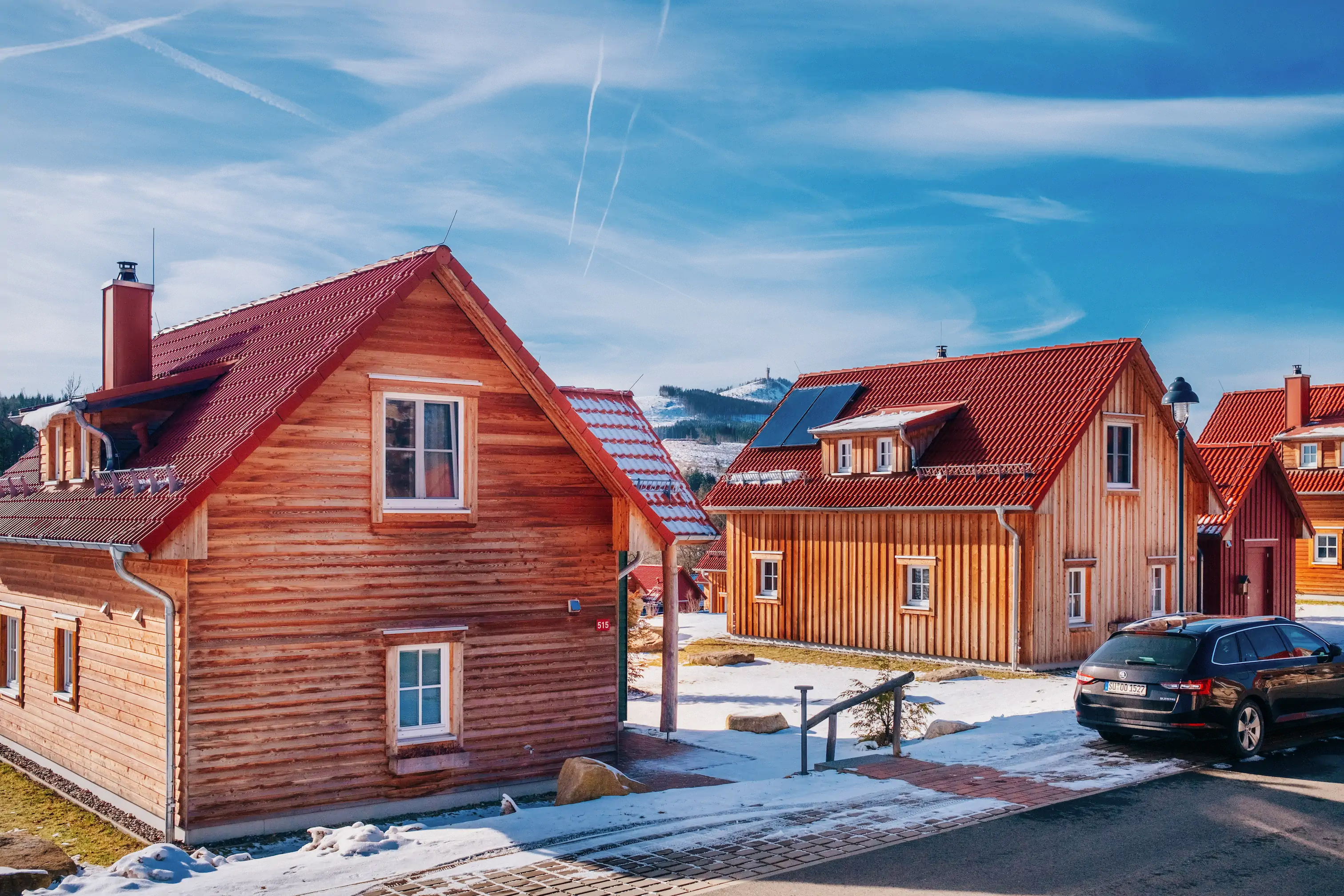 Ferienhäuser aus Holz mit roten Dächern unter blauem Himmel, geparkte Autos und Schneeflecken auf dem Boden in einer malerischen Dorfkulisse.