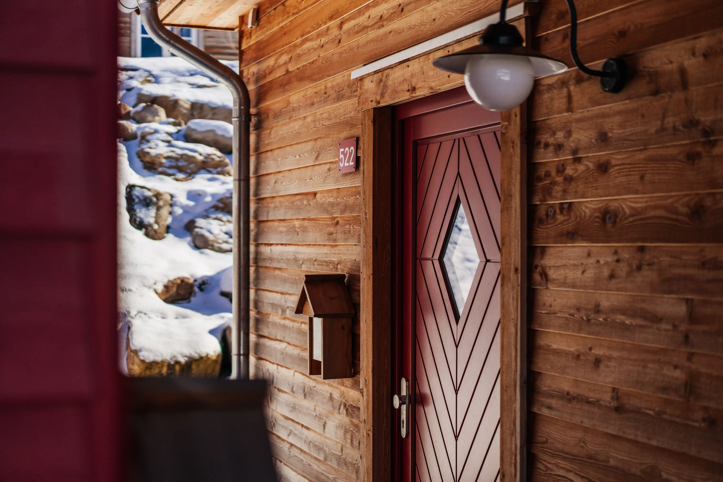 Außenansicht einer Holzhütte mit roter Tür und Rautenfenstern, einer Wandlaterne, einem kleinen Briefkasten sowie schneebedeckten Steinen im Hintergrund.
