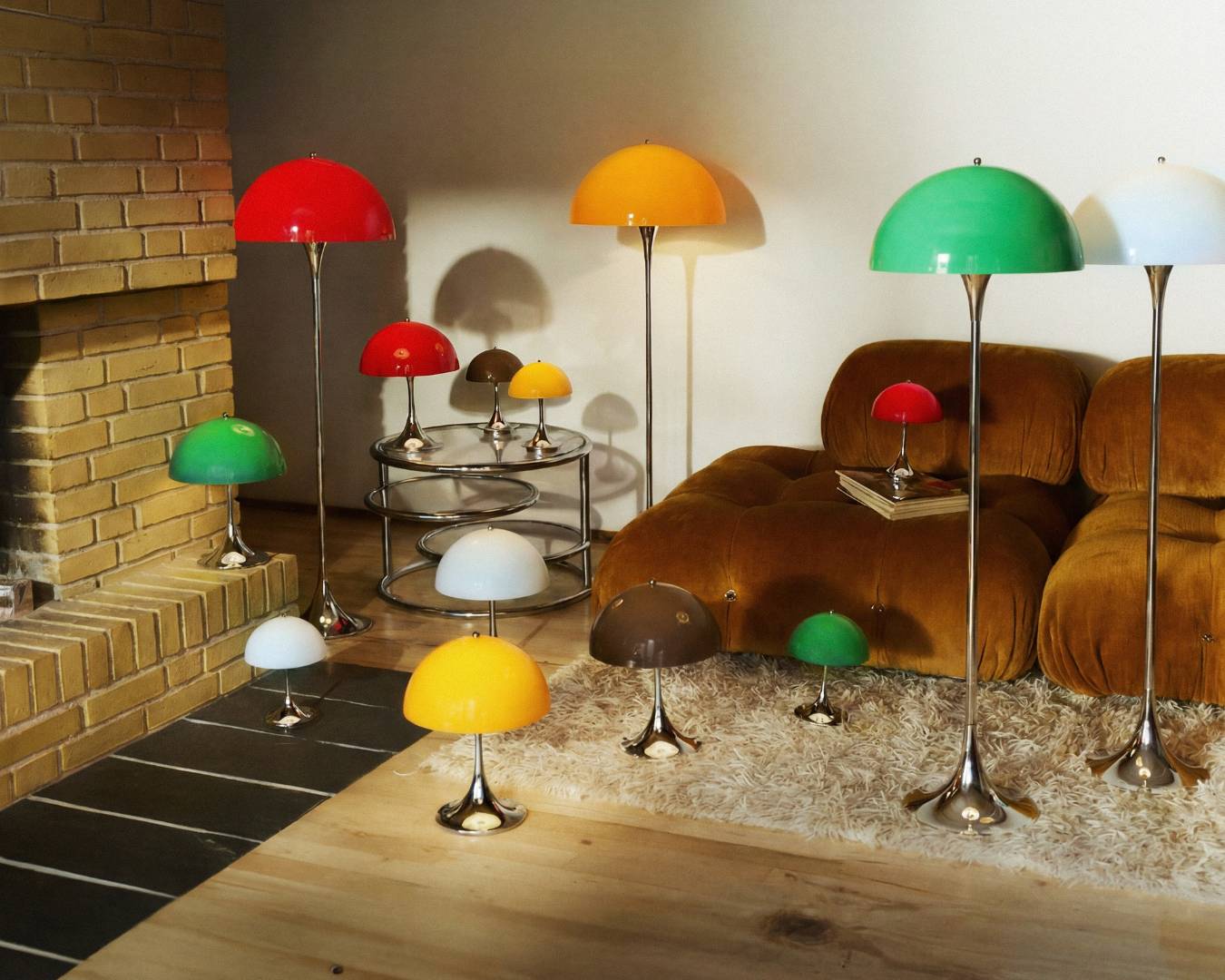 A cozy living room with colorful, dome-shaped designer lamps on the floor and table, near a brick fireplace and a plush brown sofa.