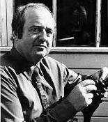Black and white portrait of a man in a shirt. He sits casually outdoors, holding something in his hands with a gentle, thoughtful expression.