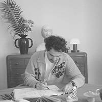 Colour image of a man with curly hair wearing a colourful sweater. He sits at a table and draws, surrounded by artistic objects and plants in a cosy environment.