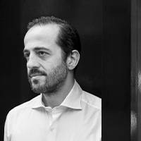 Zwart-wit afbeelding van een man met een baard en een bedachtzame blik. Hij draagt een wit shirt en staat voor een minimalistische achtergrond.