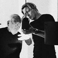 Image en noir et blanc de deux hommes en plein processus de création. L'un tient une lampe tandis que l'autre observe en détail. Tous deux sont vêtus de vêtements sombres sur un fond simple.