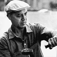 Immagine in bianco e nero di un uomo con berretto e camicia che lavora con concentrazione a un'opera d'arte, circondato da uno sfondo esterno.