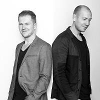 Imagem a preto e branco de dois homens, ambos com casacos informais. Estão um ao lado do outro, em frente a um fundo neutro, com uma atitude calma.