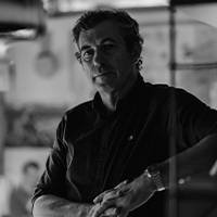 Portrait en noir et blanc d'un homme en tenue décontractée, debout dans un environnement de travail créatif en intérieur avec un faible éclairage.