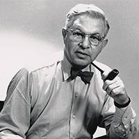 Black and white portrait of an elderly man wearing a shirt and bow tie. He holds a pipe in his hand and has a confident look with a neutral background.