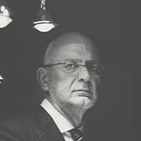 Portrait en noir et blanc d'un homme âgé, chauve, en costume-cravate. Il se tient devant des lampes suspendues à un fond sombre.