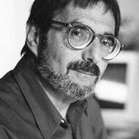 Imagen en blanco y negro de un hombre con gafas y una cálida sonrisa. Tiene el pelo rubio y lleva un jersey ante un fondo sencillo.