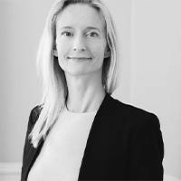 Black and white image of a woman with long, blonde hair and a friendly smile. She is dressed in a simple blouse and stands in front of a bright background.