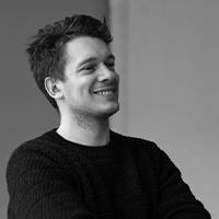 Imagem a preto e branco de um jovem com cabelo curto e um rosto sorridente. Veste uma camisola e está sentado casualmente em frente a um fundo neutro.