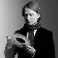 Retrato a preto e branco de um homem com cabelo semi-longo, vestindo um blazer e um cachecol. Segura um objeto artístico com um olhar concentrado.