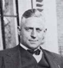 Immagine in bianco e nero di un uomo in abito e papillon. Ha uno sguardo acuto e i capelli corti e raccolti su uno sfondo esterno.