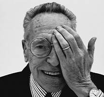 Zwart-wit portret van een oudere man met bril en glimlach, die één oog bedekt met zijn hand. Hij is gekleed in een klassiek overhemd en blazer.