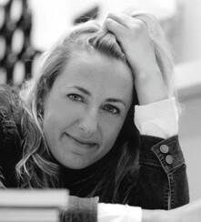 Image en noir et blanc d'une femme aux cheveux mi-longs se soutenant la tête d'une main. Elle sourit avec décontraction devant un fond créatif.