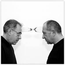 Mustavalkoinen kuva kahdesta vastakkain seisovasta miehestä, jotka on erotettu toisistaan silmälaseilla. He ovat pukeutuneet tummiin villapaitoihin ja minimalistiseen taustaan.