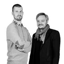 Zwart-wit portret van twee mannen, de ene staat nonchalant met zijn armen over elkaar en de andere draagt een sjaal. Beiden staan voor een witte achtergrond.