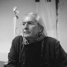 Černobílý portrét staršího muže s polodlouhými vlasy a brýlemi. Má na sobě šálu a tmavou bundu a stojí v kreativním pracovním prostředí.