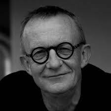 Black and white image of a man with round glasses and a friendly smile. He has short hair and sits in a relaxed pose in front of a neutral background.