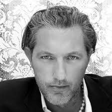 Fekete-fehér portré egy elegáns stílusú, feszült tekintetű férfiról. Félhosszú haja van, és részletes háttérmintás ing és blézer van rajta.