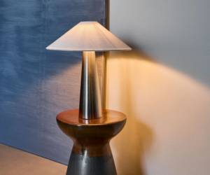 Tischleuchte mit konischem Schirm und Chromfuß auf einem Holzbeistelltisch vor blauer Wand