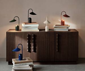 Mai multe lămpi de masă colorate așezate pe o măsuță din lemn închis la culoare, deasupra unor teancuri de cărți.