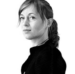 Portrait en noir et blanc d'une femme aux cheveux coiffés et au regard calme. Elle porte un pull noir et le fond est neutre.