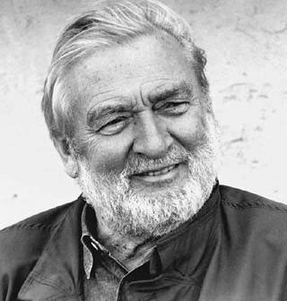 Czarno-biały portret uśmiechniętego, brodatego mężczyzny o zrelaksowanym wyrazie twarzy. Ma zaczesane do tyłu włosy i nosi kurtkę. Tło jest neutralne i niezakłócone, podkreślając charakter twarzy.