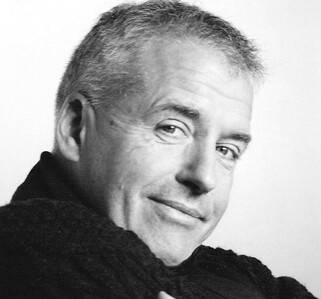 Imagem a preto e branco de um homem mais velho com um sorriso torto e cabelo curto. Está vestido com uma camisola e olha amigavelmente para a câmara.