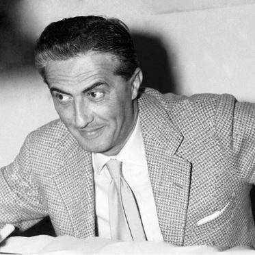 Black and white image of a man in a suit and tie. He has short hair and a crooked smile, sitting at a table with a simple background.