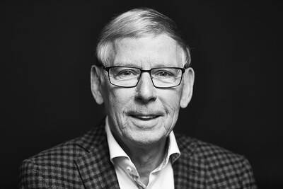 Černobílý portrét muže v obleku a košili. Přátelsky se usmívá a nosí brýle, zatímco pozadí je tmavé a jednoduché.