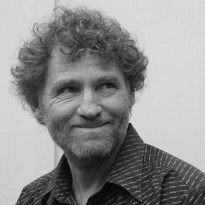 Mustavalkoinen muotokuva miehestä, jolla on kiharat hiukset ja parta. Hän hymyilee helposti, ja hänen käytöksensä on rento yksinkertaista taustaa vasten.