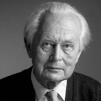 Čiernobiely obrázok staršieho muža s bielymi vlasmi, ktorý má na sebe kravatu a sveter. Má zamyslený a vážny výraz na tmavom pozadí.