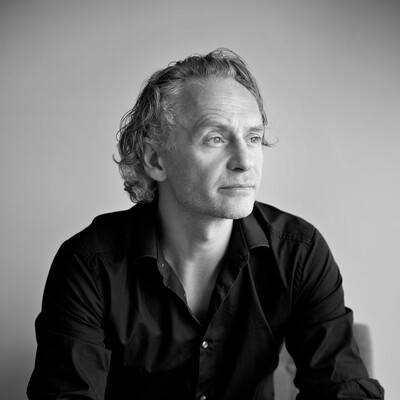 Ritratto in bianco e nero di un uomo con capelli ricci semilunghi e sguardo pensieroso. Indossa una camicia nera ed è seduto davanti a uno sfondo neutro.