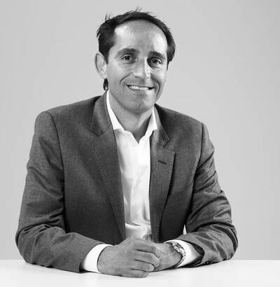 Immagine in bianco e nero di un uomo con i capelli corti, che indossa una giacca e una camicia. Siede sorridente a un tavolo con un atteggiamento rilassato e professionale.