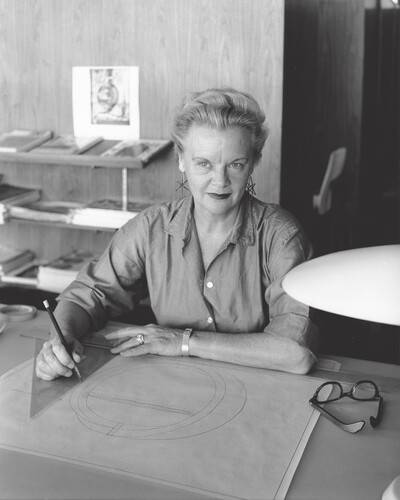 Svart-hvitt portrett av en kvinne ved arbeidsbordet sitt. Hun tegner og er omgitt av verktøy og en lampe, med bøker på hyller i bakgrunnen.