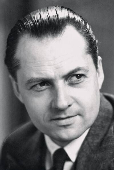 Portrait vintage en noir et blanc d'un homme aux cheveux gominés et à l'air sérieux. Il porte un costume et une cravate, sur un fond simple.