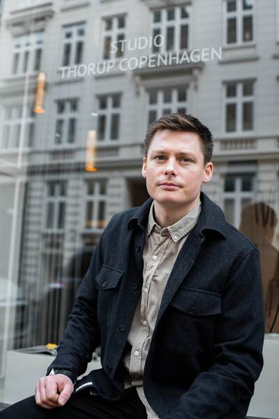 Mężczyzna siedzi przed oknem z napisem "Studio Thorup Copenhagen" w tle. Ubrany jest w ciemną kurtkę i pewnie spogląda w kamerę