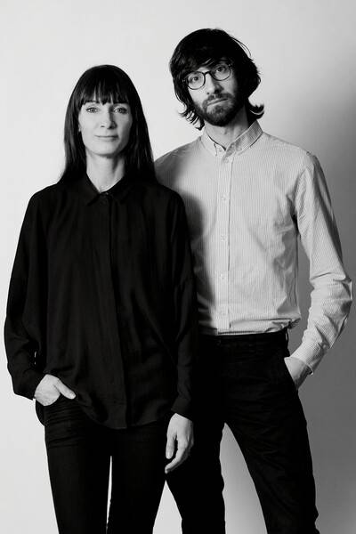 Black and white portrait of a woman and a man. The woman is dressed in a dark shirt and stands casually with her hand in her pocket. The man wears glasses and a light-coloured shirt, both standing in front of a neutral background.