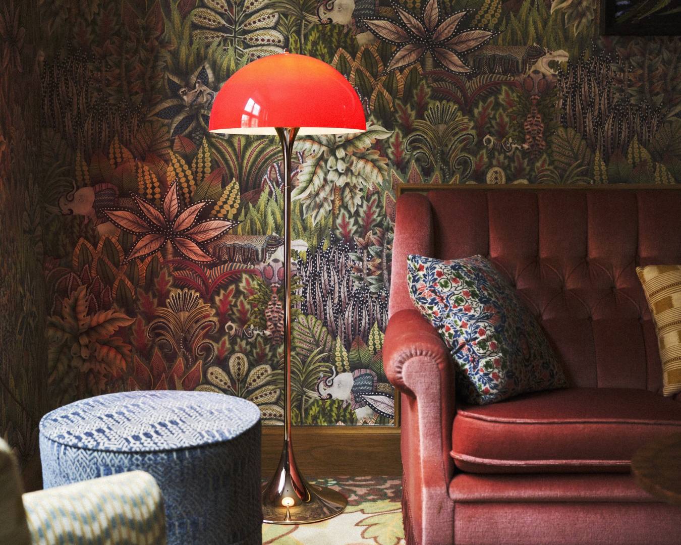 Cozy interior with a retro red floor lamp, patterned wallpaper, a textured blue ottoman, and a plush red sofa accented by a floral pillow.