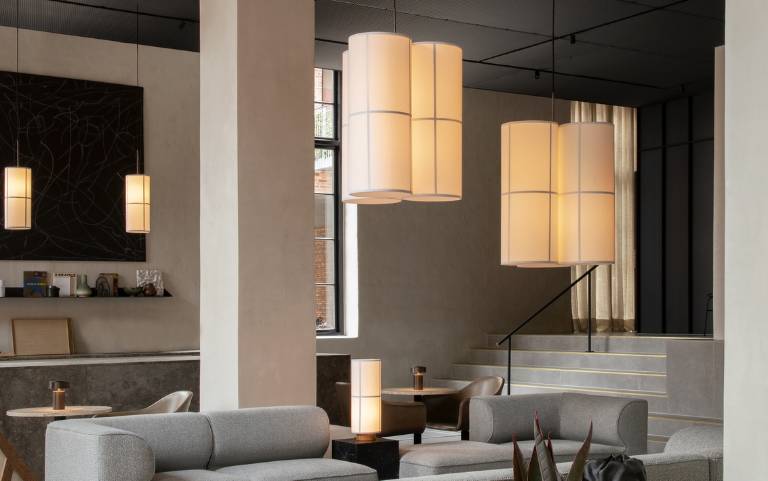 super! Einen Alttext zu dem Bild  Mehrere Hashira Pendelleuchten und eine Hashira Tischleuchte von Audo Copenhagen mit warm leuchtenden Textilschirmen in einem Lounge-Bereich mit grauen Sofas.