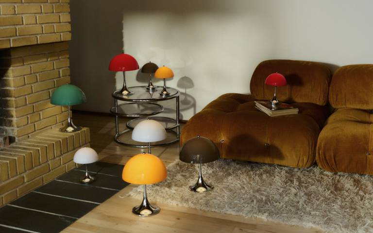 Flere Panthella bordlamper i forskellige farver står i et hyggeligt rum med en brun sofa, et tæppe og en muret pejs. Lamperne skaber varme, bløde lysglimt på gulvet,og sofaen.