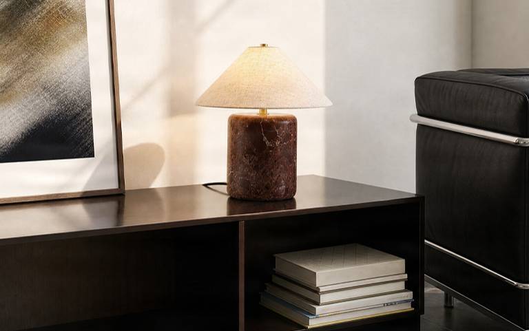 Lampa stołowa Nola z czerwoną marmurową podstawą i lnianym kloszem na ciemnej komodzie w promieniach słońca