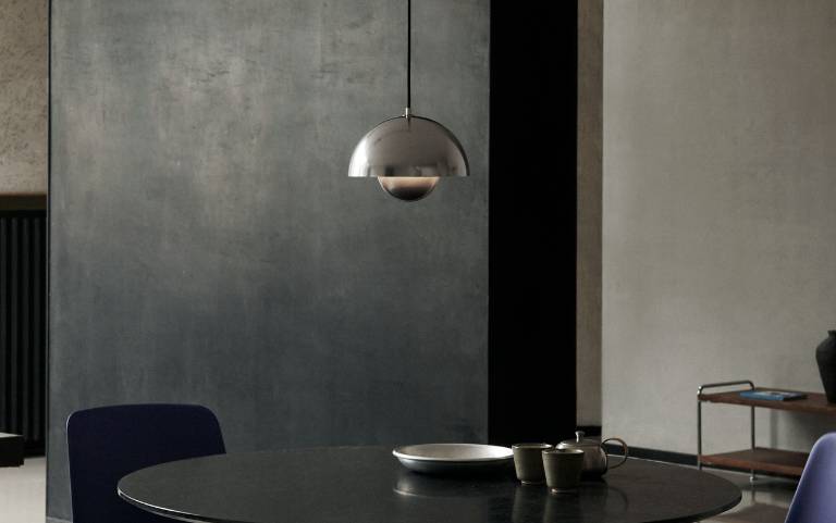Lampa wisząca &Tradition Flowerpot VP1 z cyny zawieszona nad ciemnym, okrągłym stołem jadalnym z ceramicznymi naczyniami, na tle szarej betonowej ściany.