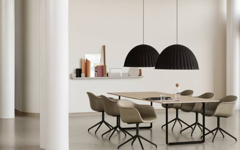 Zwei schwarze Under the Bell Pendelleuchten von Muuto über einem Eichenholztisch mit hellbeigen Fiber Armchairs in einem hellen, offenen Raum.