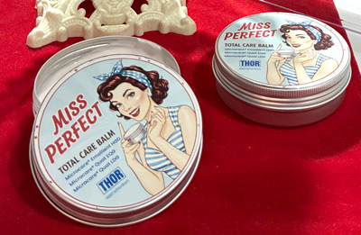 Tin cans of "Miss Perfect" total care balm with vintage-style illustrations of a woman on a red fabric background.