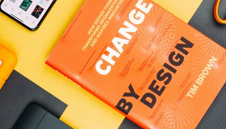 Sehr komponiertes Foto eines Schreibtisches mit verschiedenen Büroutensielien, alles drapiert rund um das Buch "Change by Design" von Tim Brown
