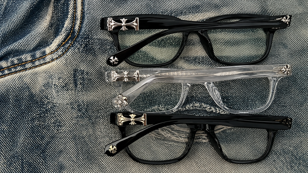 Three pairs of glasses, two black and one clear, with cross designs on the arms, placed on a blue denim fabric background.